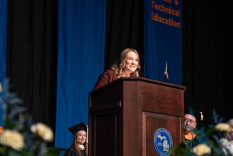 Beth LaForest stands at a podium delivering the 2025 commencement address at North Central Michigan College, with faculty and graduates seated behind her.