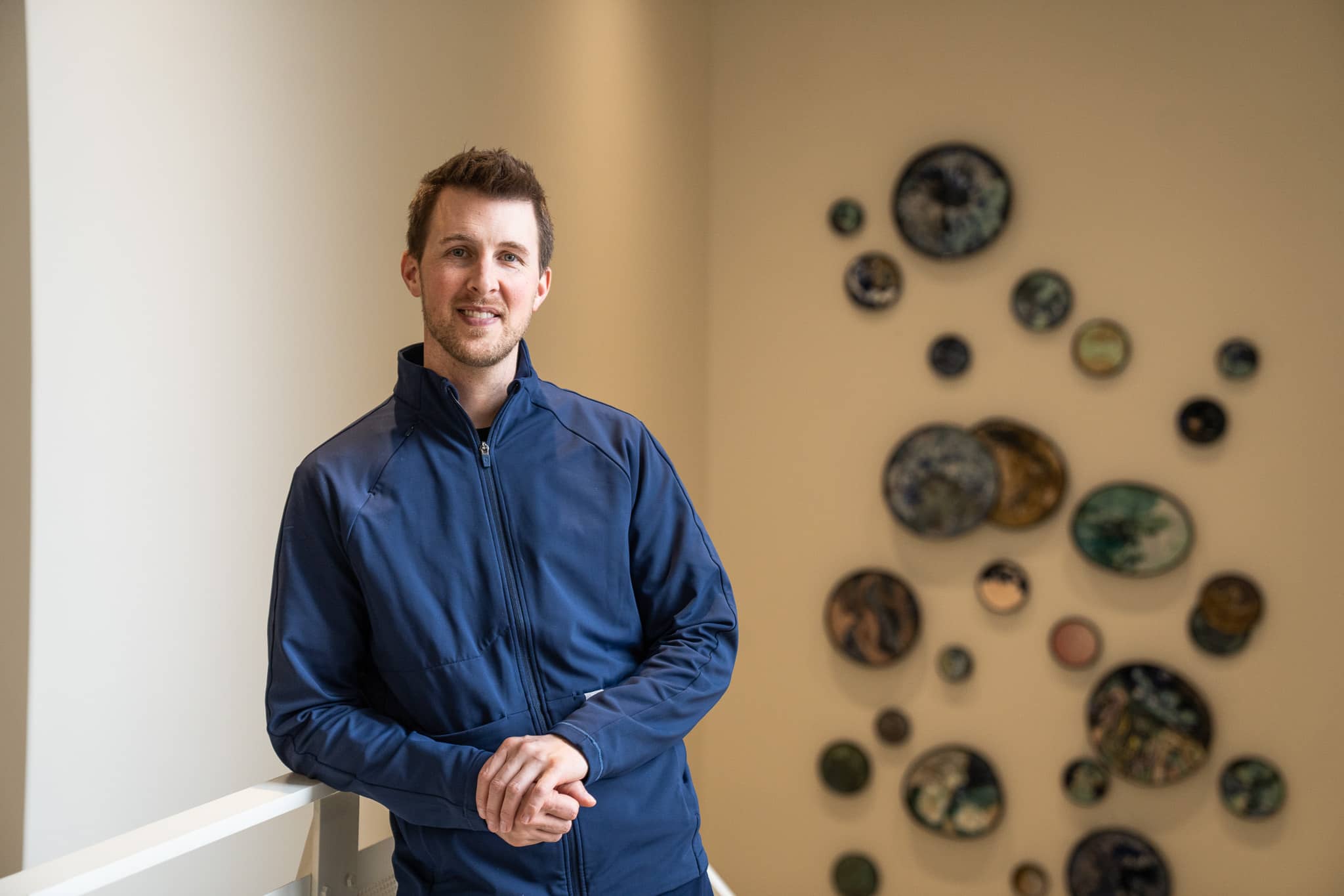 Caleb Migda, a North Central Michigan College nursing graduate, stands indoors near a wall display of ceramic art pieces.