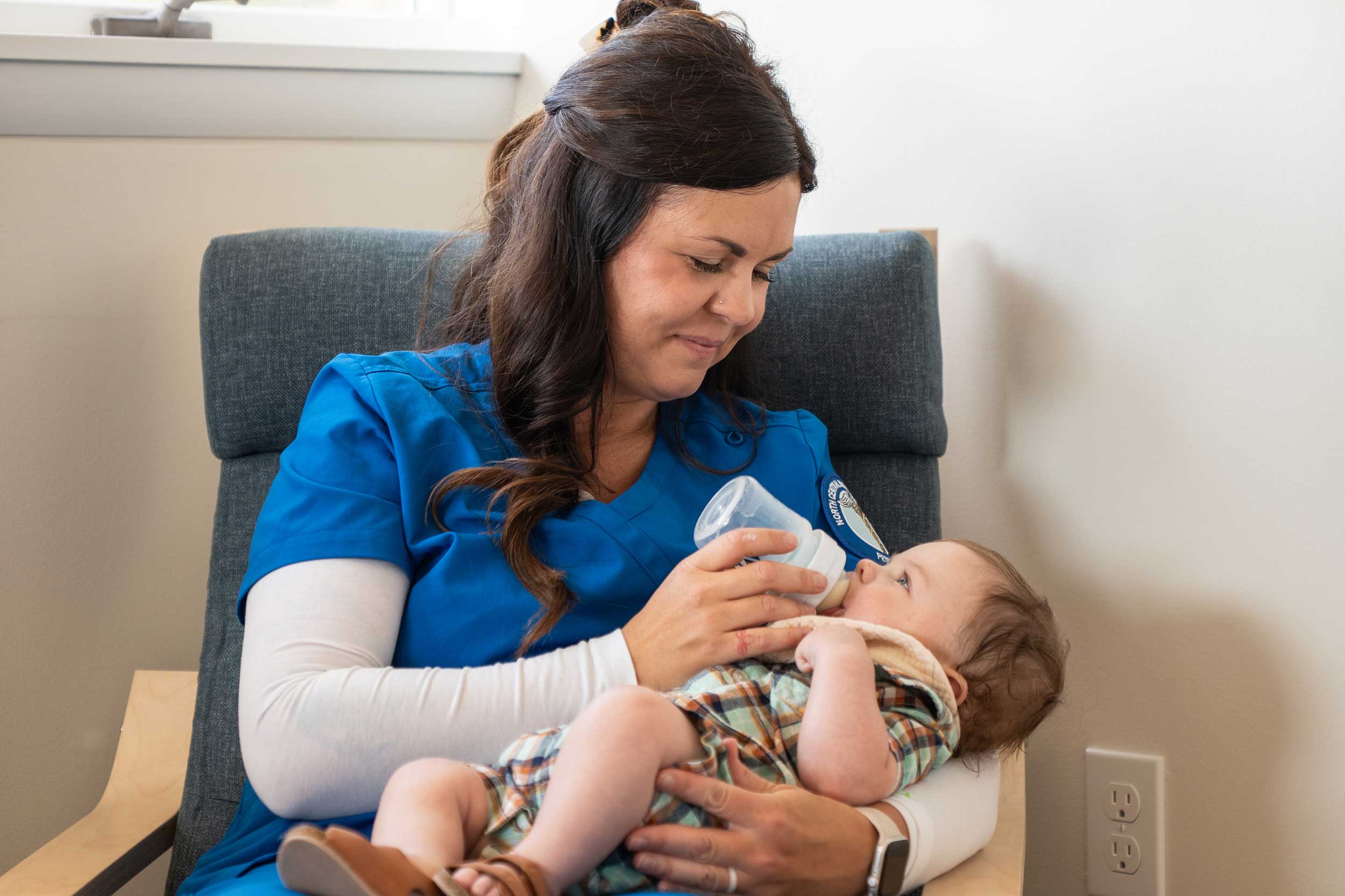 NCMC nursing student wearing blue scrubs and feeding her baby a bottle.