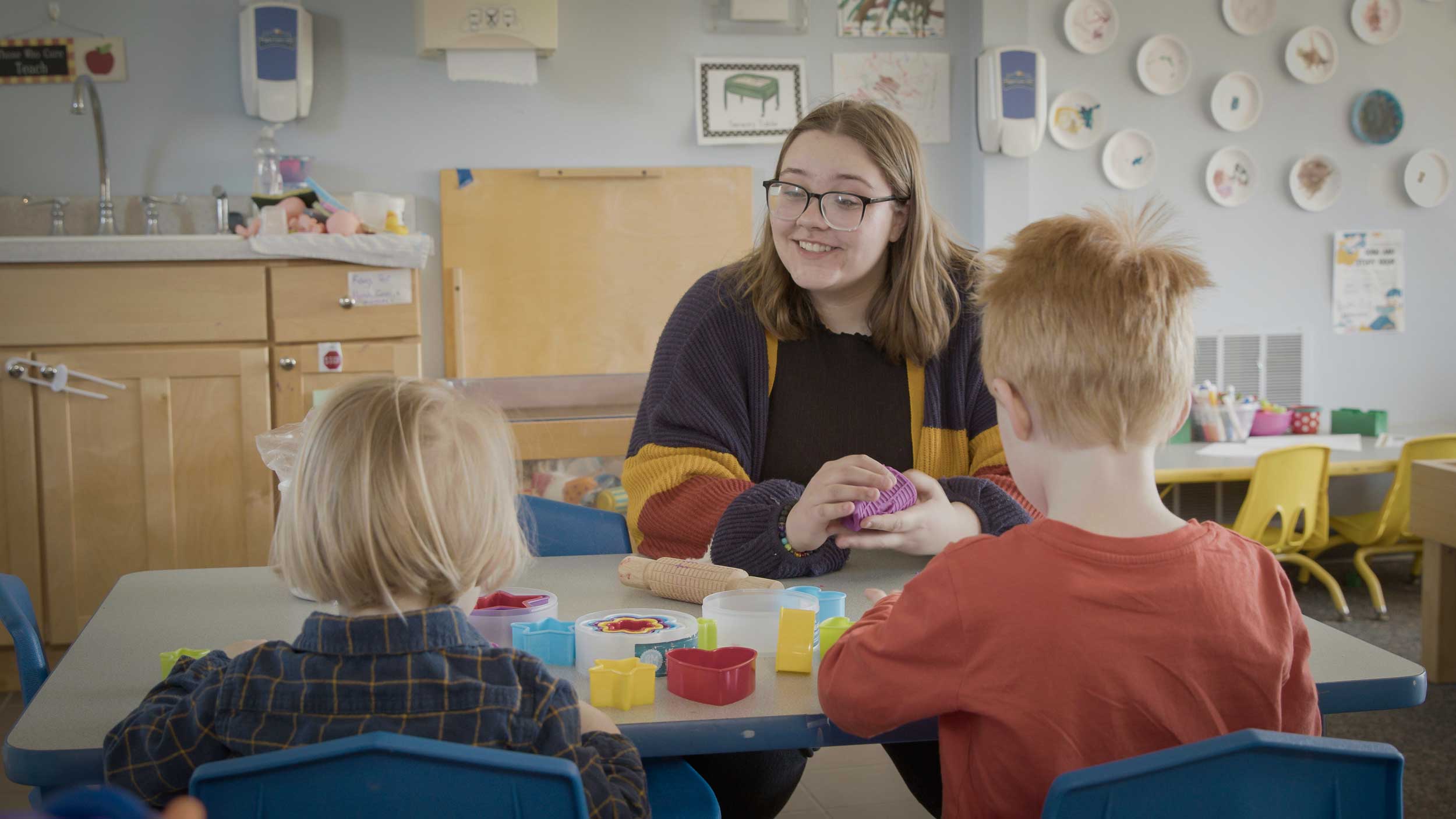 Kaitlyn Hammerle, a North Central Michigan College Early Childhood Education alumna, smiles while interacting with two young children in a classroom setting, engaging them in a hands-on activity with colorful toys.