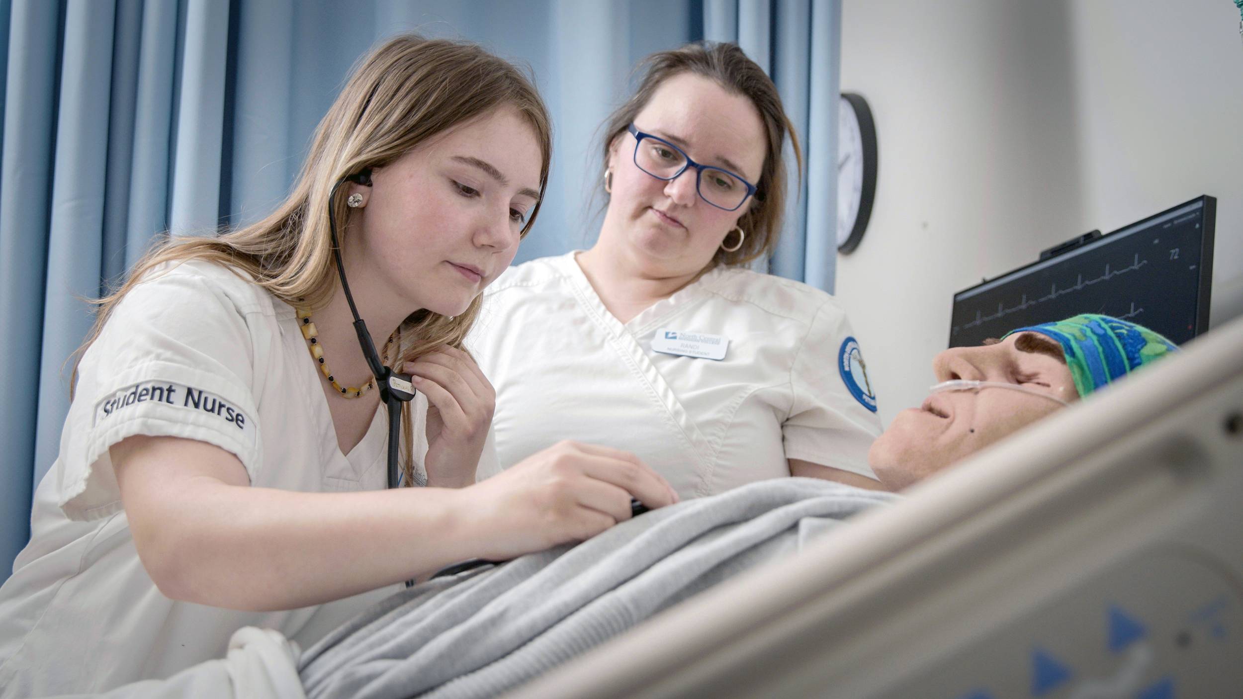 A young student nurse listens to a patient’s chest with a stethoscope while another nurse, who appears to be supervising, looks on. Both are wearing North Central Michigan College nursing uniforms.