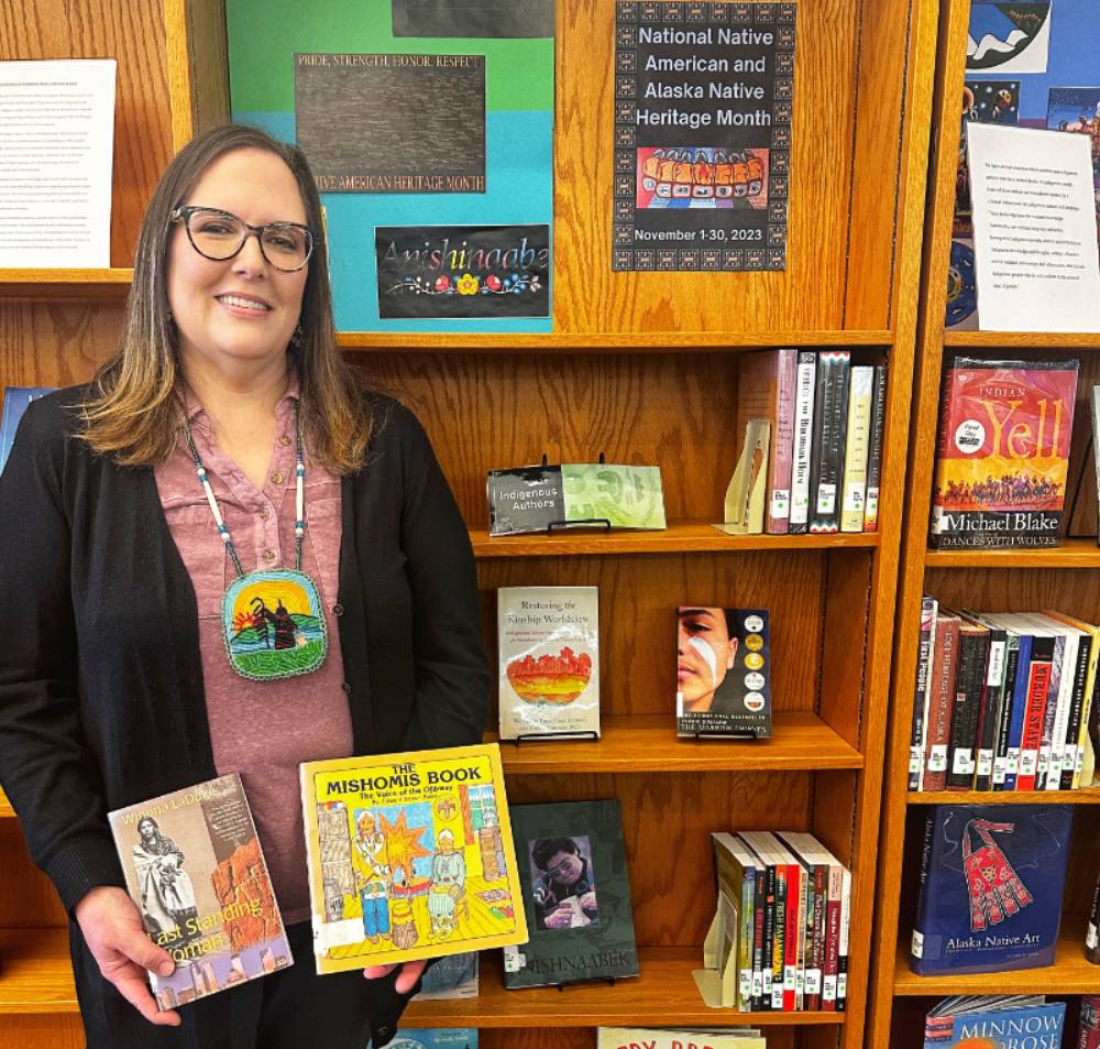 Beth Fisher-Polasky, Library Specialist at North Central Michigan College, holding books from the reorganized Indigenous collection, standing in front of a library display celebrating Native American Heritage Month.