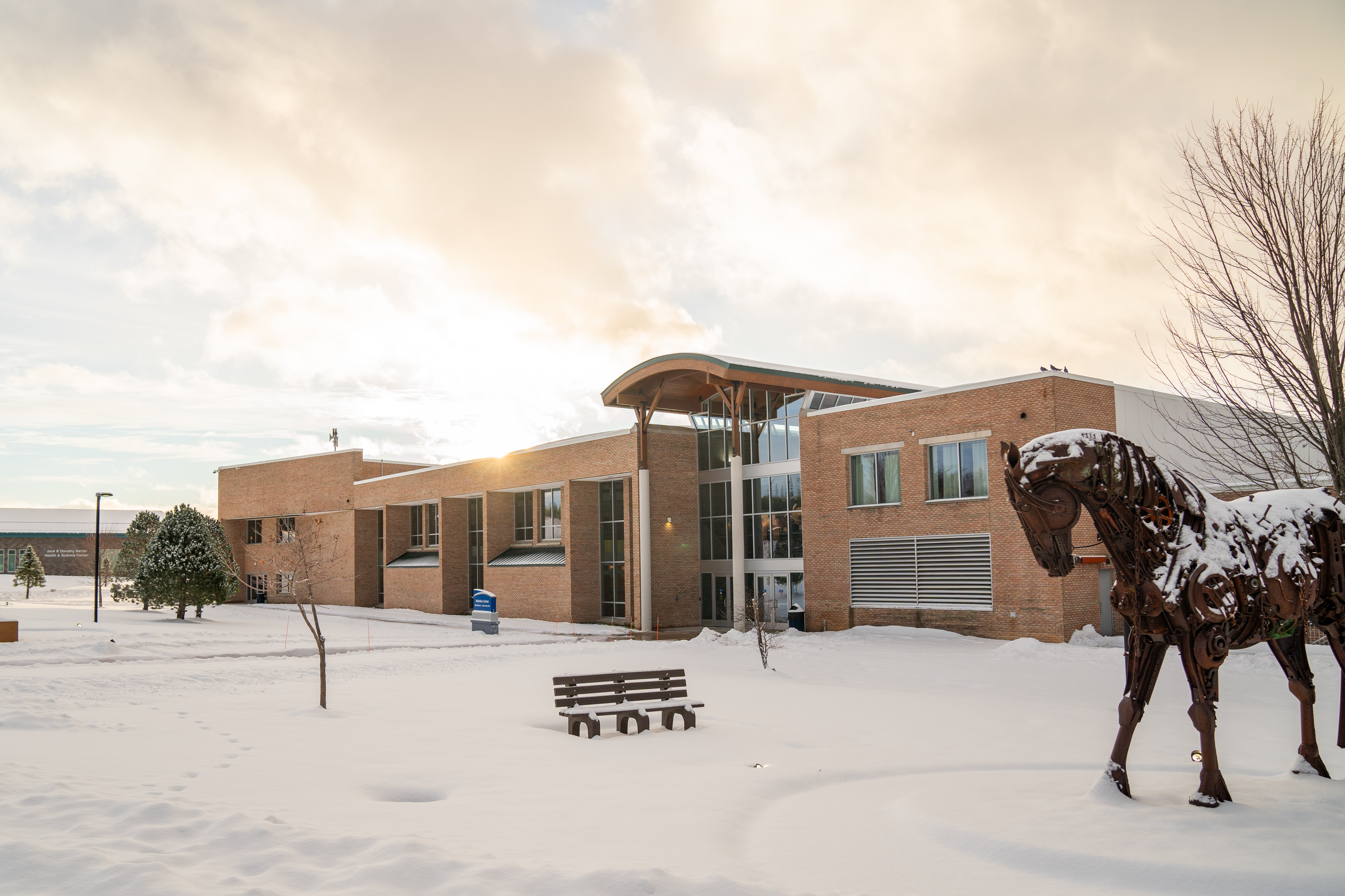 Photograph of North Central Michigan College’s Athletic Center in Petoskey, Michigan, showcasing a snowy campus sunrise with sculptures and the Jack and Dorothy Harris Health Education and Science Center in the background