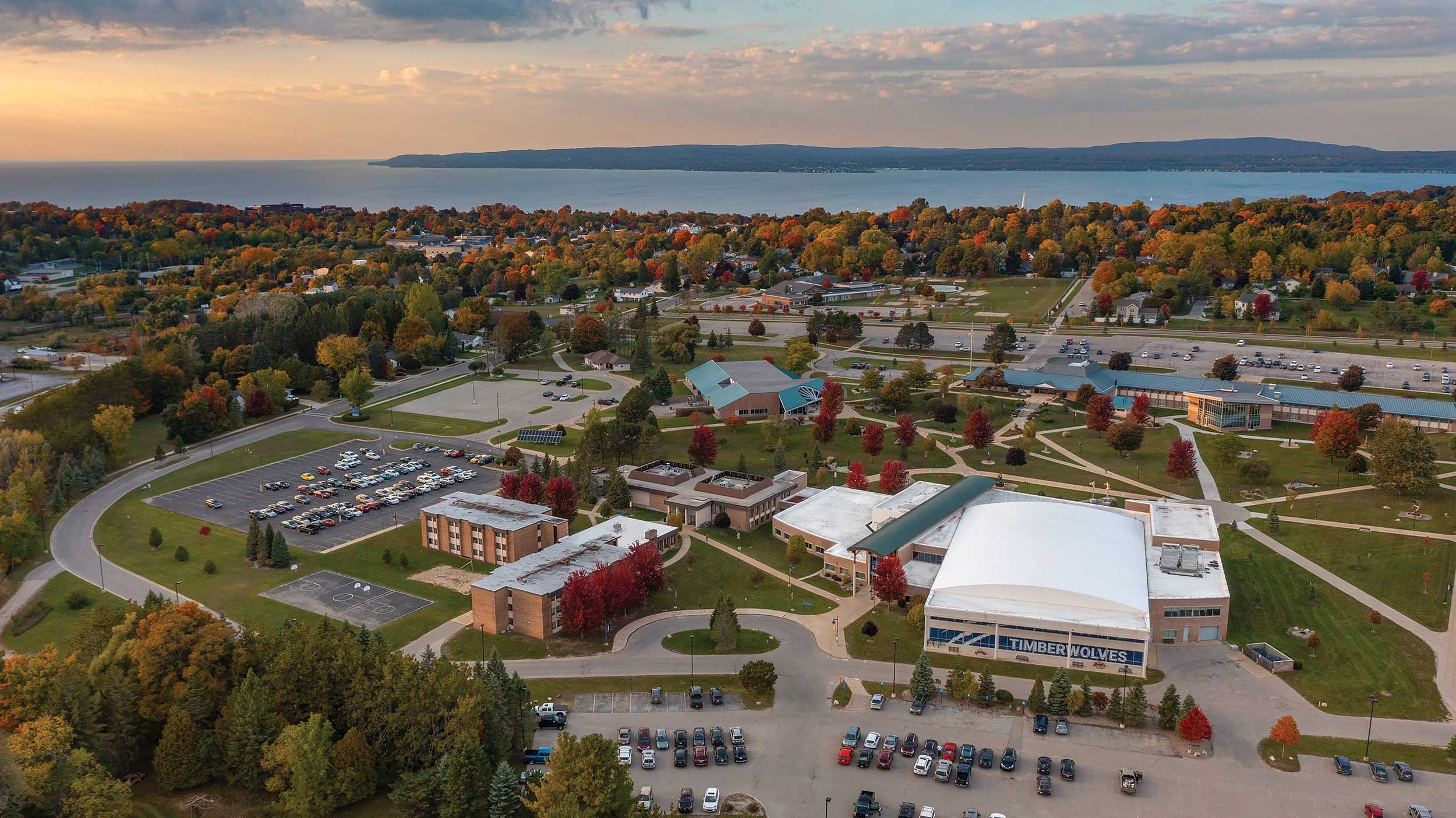 Aerial view of North Central Michigan College's campus in autumn, with trees surrounding buildings and Lake Michigan in the background