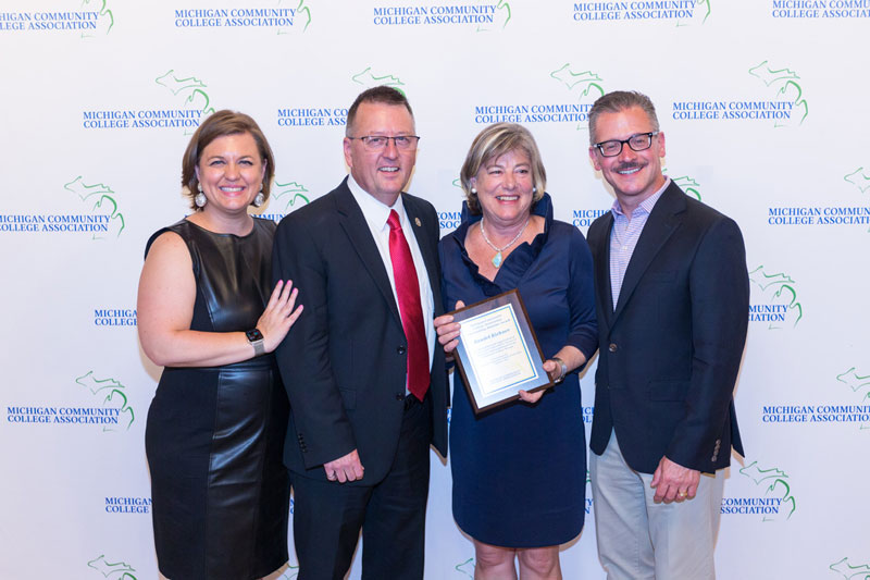 Randel Richner, second from right, receives the Outstanding Community College Alumnus Award from the Michigan Community College Association. She is pictured with three colleagues against a backdrop displaying the MCCA logo.